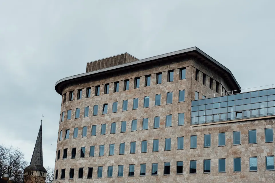 Modernes Gebäude mit Glasfenstern und nahegelegenem Kirchturm vor einem bewölkten Himmel.